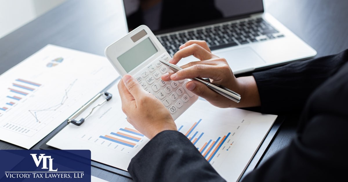 Hands working a calculator over financial charts beside a laptop, Victory Tax Lawyers